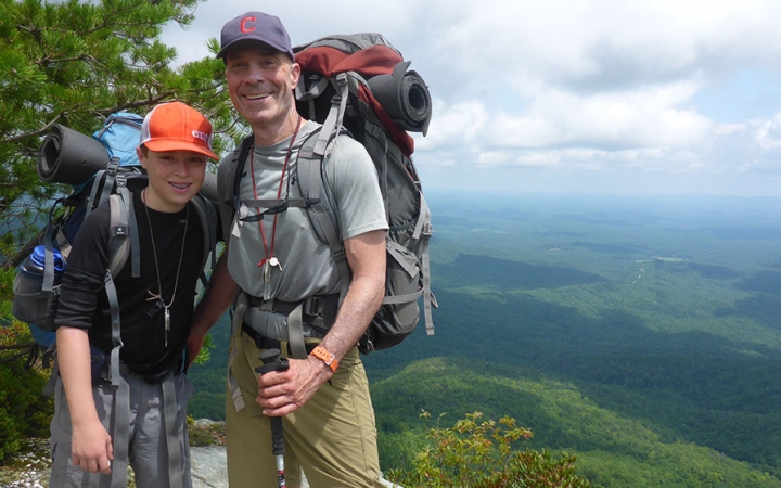 A parent and child smile for a photo on an overlook high above a green mountainous area. Both are wearing backpacks.